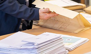 Ablage und Dokumentenprüfung von Bauakten Eine Person sortiert Bauakten und Baupläne an einem Schreibtisch. Im Vordergrund liegt ein Stapel von Dokumenten, während im Hintergrund weitere Aktenmappen bereitliegen. Das Bild vermittelt den Arbeitsalltag mit Papierdokumenten und zeigt den sorgfältigen Umgang mit wichtigen Bau- und Verwaltungsakten.