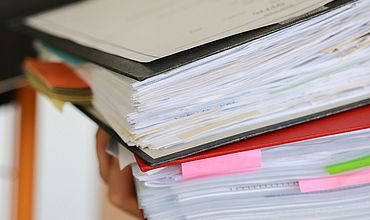 Papierflut im Büroalltag Ein großer Stapel Aktenordner und Papierdokumente wird von einer Hand getragen. Die Akten sind mit bunten Markierungen versehen, was auf einen arbeitsintensiven und gut organisierten Bürotag hinweist. Das Bild vermittelt den Eindruck von hoher Arbeitsbelastung und der Notwendigkeit, viele wichtige Unterlagen zu verwalten.